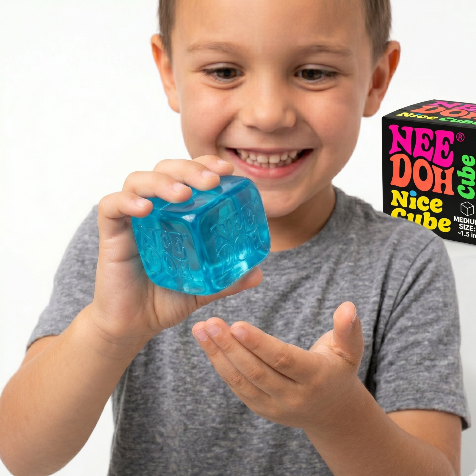 A smiling young boy playfully squishing a blue translucent NeeDoh Nice Cube in his hands, with the official Schylling Nice Cube box visible in the background.